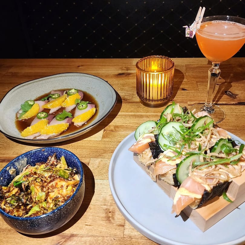 Assortment of Japanese dishes including sashimi, sushi, and a side dish, with a cocktail and candle.
