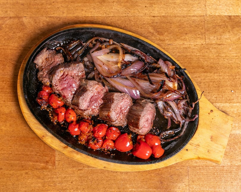 Sizzling steak pieces, roasted red onions, and cherry tomatoes on a black cast iron platter.