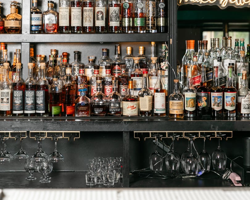 A well-stocked bar shelf displays numerous bottles of liquor, with hanging glasses below.