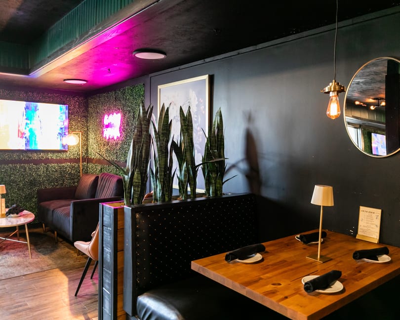 A restaurant interior with a booth, wooden table, modern lighting, and a lounge area with a TV.