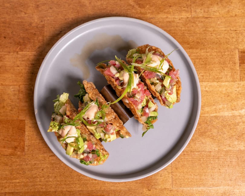 Four tuna poke tacos with avocado, sauce, and scallions on a gray plate.