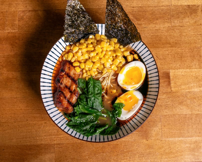 Ramen bowl with grilled pork, corn, spinach, soft-boiled eggs, and nori on a wooden table.
