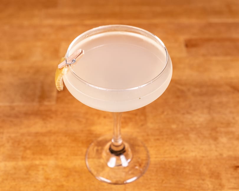 Light-colored cocktail in a coupe glass, garnished with a lemon peel held by a tiny clothespin.