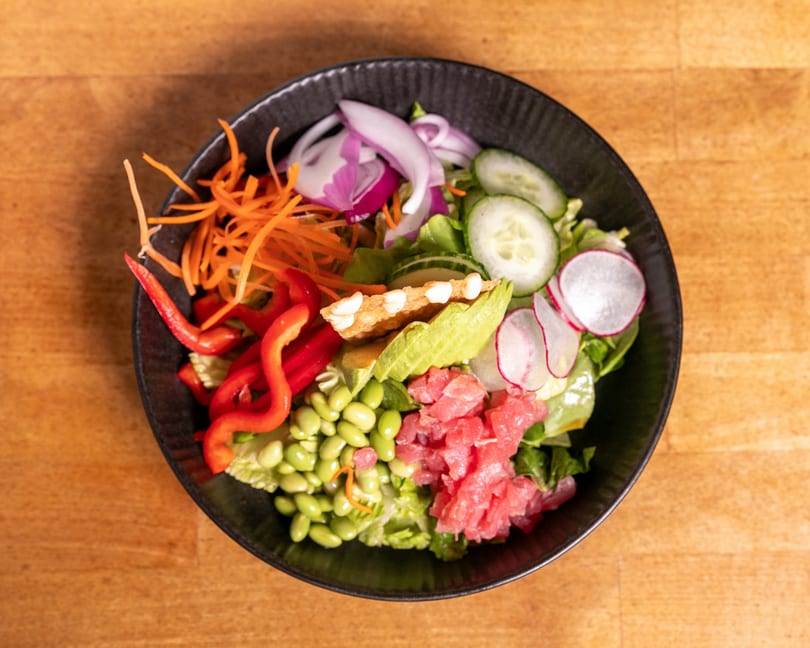Overhead view of a colorful salad with tuna, avocado, edamame, and vegetables in a black bowl.
