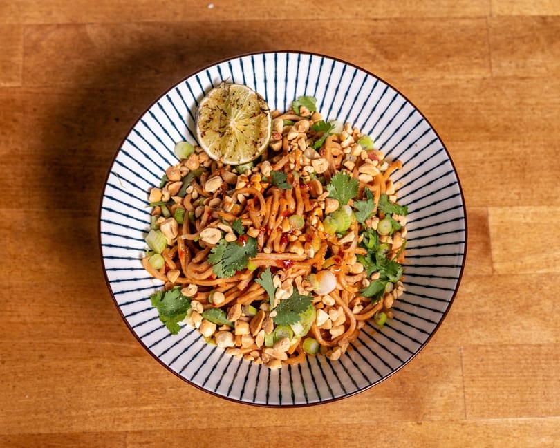 A bowl of noodles with peanuts, cilantro, green onions, and a grilled lime wedge.