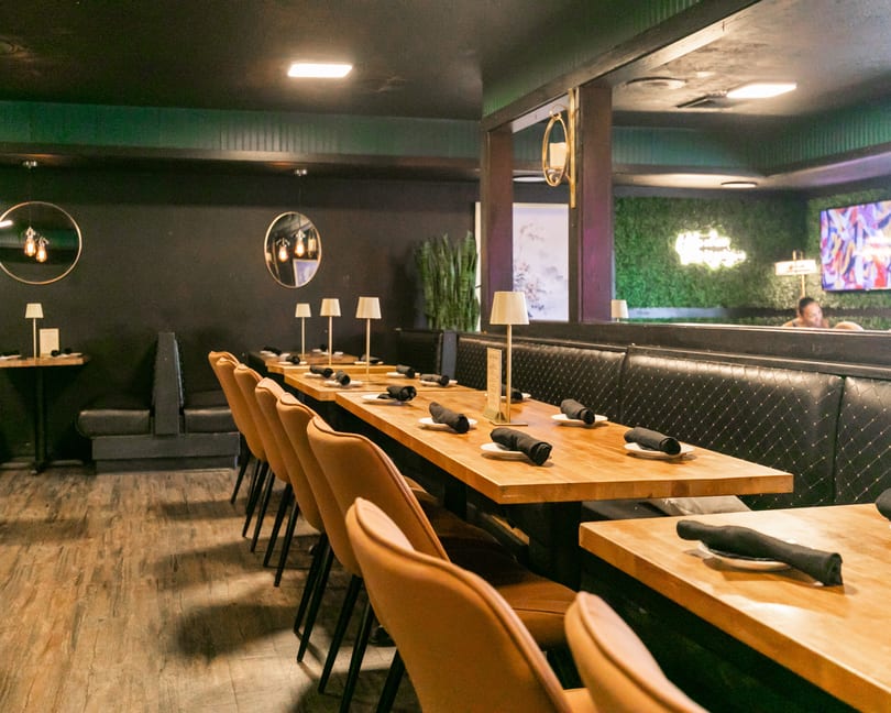 Restaurant interior with long wooden tables, brown chairs, black booths, and dark green walls.
