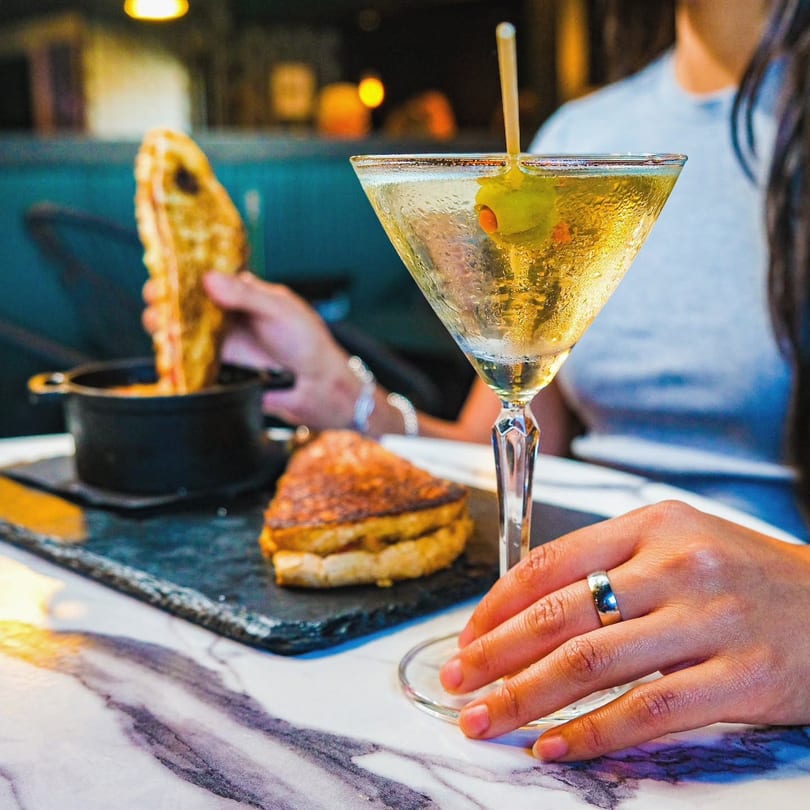 A person holds a martini with an olive, next to a grilled cheese sandwich and tomato soup.