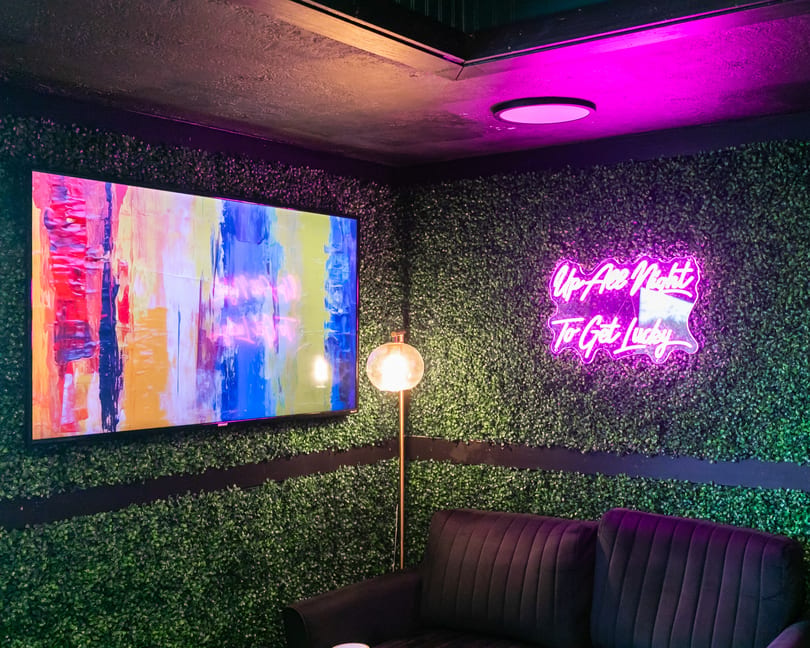 A lounge area with a green hedge wall, a colorful abstract TV, and a neon sign reading "Up All Night To Get Lucky".