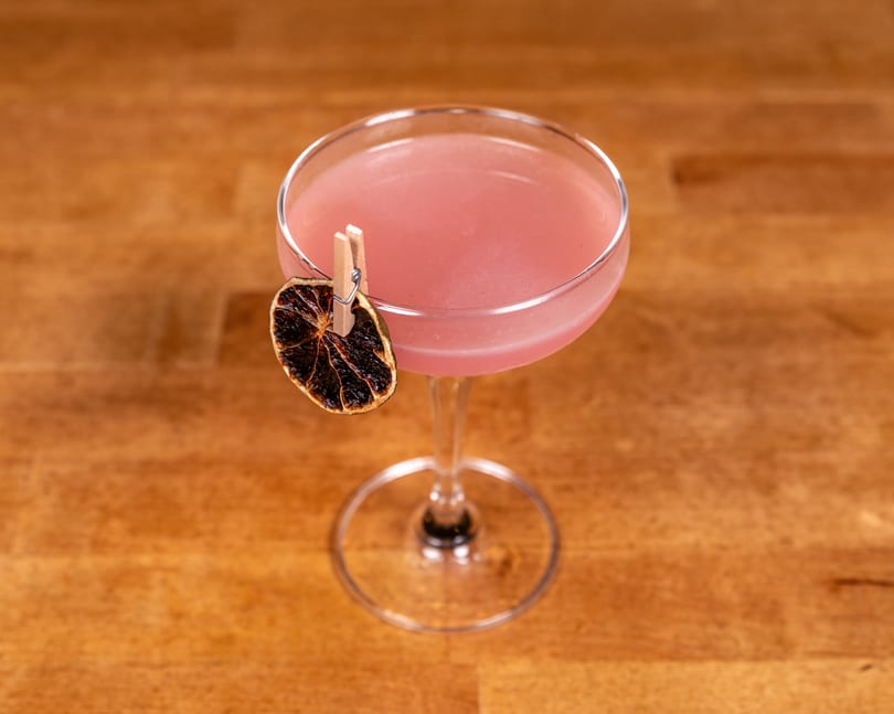 Pink cocktail in a coupe glass, garnished with a dried citrus slice on a wooden table.