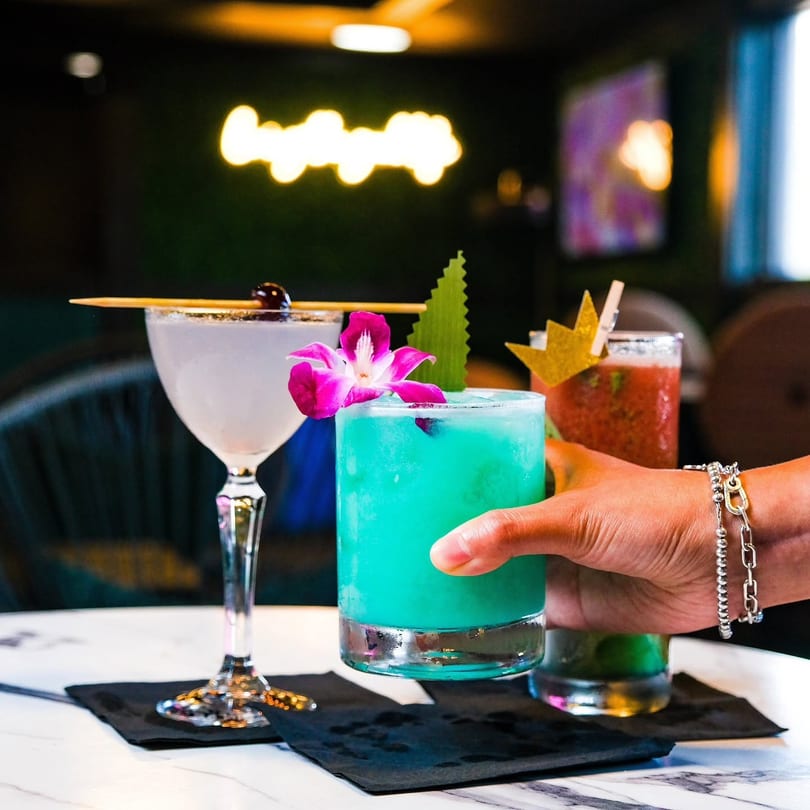Three colorful cocktails on a table, one held by a hand, with a neon sign in the background.