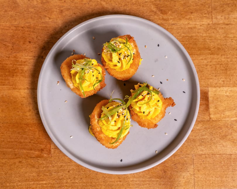 Four crispy deviled eggs topped with yellow filling, green onions, and sesame seeds on a gray plate.