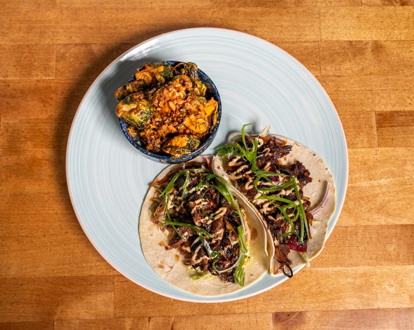 Two tacos with meat, onions, and green garnish, alongside a bowl of roasted Brussels sprouts.