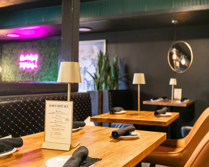 A restaurant interior with wooden tables, black chairs, a neon sign, and table lamps.
